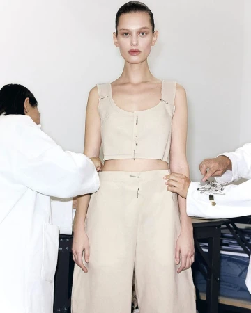 A model poses in a cropped top and wide-legged pants while being fitted by two unseen tailors in white lab coats.