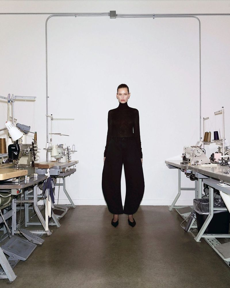 A slender adult female stands centered in a fashion-forward outfit featuring a black turtleneck and balloon pants, set in a workshop with sewing machines.