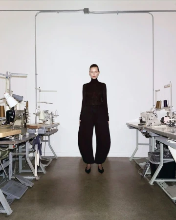A slender adult female stands centered in a fashion-forward outfit featuring a black turtleneck and balloon pants, set in a workshop with sewing machines.