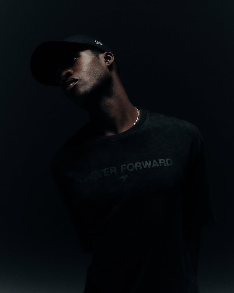 Portrait of a Black male model in a black cap and black sweatshirt, against a dark grey studio backdrop, looking up into low light.