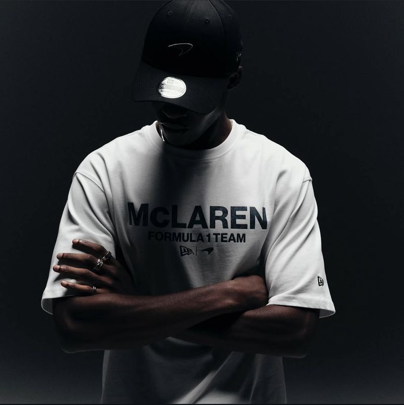 Man in McLaren Formula 1 Team t-shirt and black cap with arms crossed in a studio setting against dark backdrop.