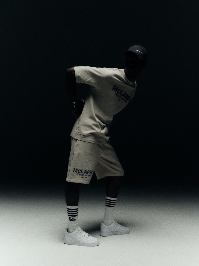 Man poses in McLaughlin Formula 1 racing t-shirt and shorts, wearing sneakers and striped socks, standing in a studio with dark backdrop.