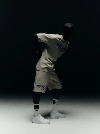 Man poses in McLaughlin Formula 1 racing t-shirt and shorts, wearing sneakers and striped socks, standing in a studio with dark backdrop.