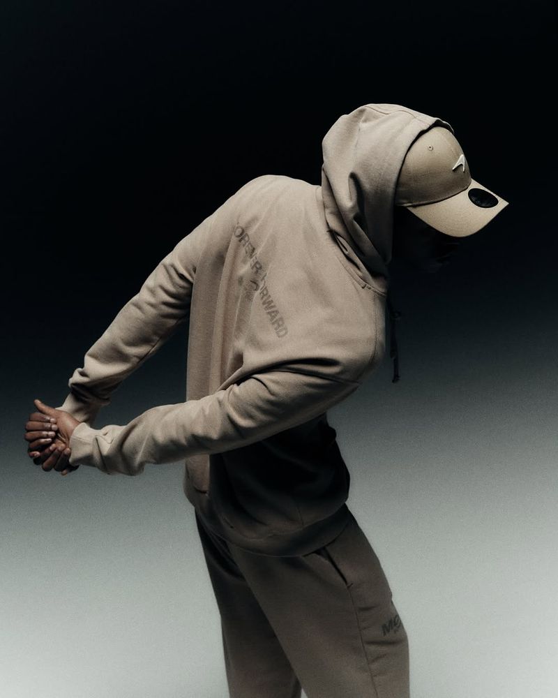 Person in a tan hooded sweatshirt and baseball cap posing against a gradient gray and black background in a fashion-style photograph.