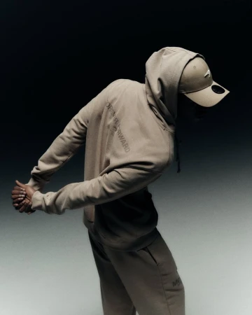 Person in a tan hooded sweatshirt and baseball cap posing against a gradient gray and black background in a fashion-style photograph.