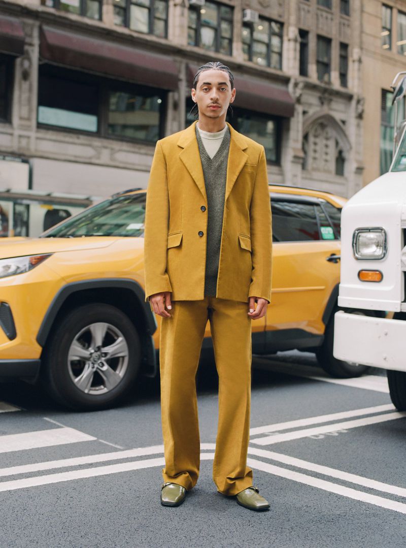 A man in a yellow suit stands on a city street near a yellow taxi and a white truck, creating a modern, urban fashion portrait.