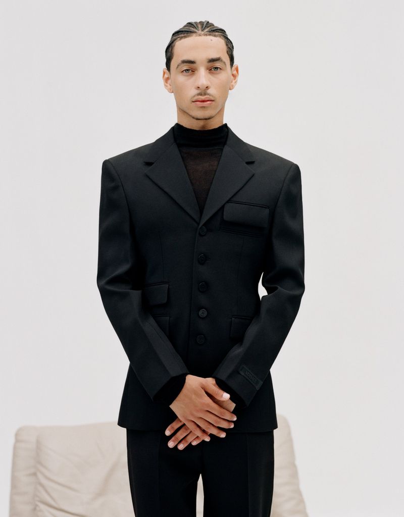 Portrait of a handsome adult male with braids, wearing a formal black suit with padded shoulders, posing in front of a white background.