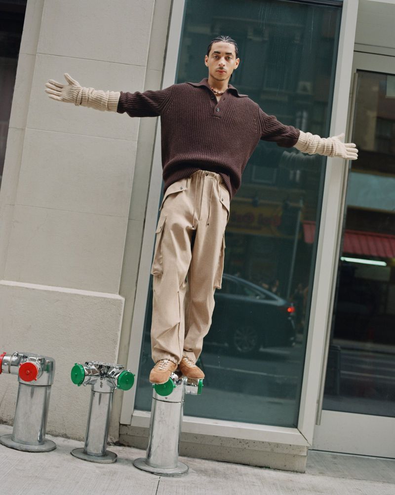 A stylish adult male stands atop a fire hydrant on an urban street, wearing brown knitwear, beige cargo pants, and hand warmers.