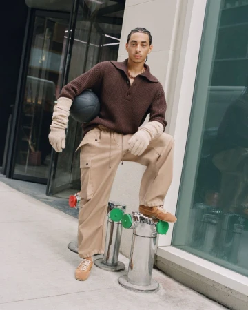 A young adult male poses outdoors in a brown sweater and beige pants, holding a basketball. He stands next to metal bollards.