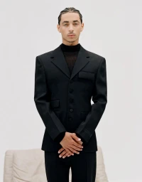 Julez Smith wearing a tailored black suit jacket over a dark turtleneck against a plain white studio backdrop.