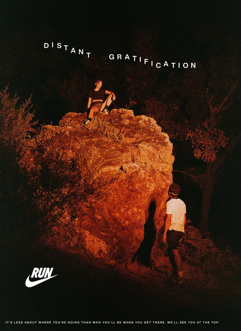 Two people in a Nike advertisement. One person stands at the base of a rock formation while the other sits at the top against a dark outdoor background.
