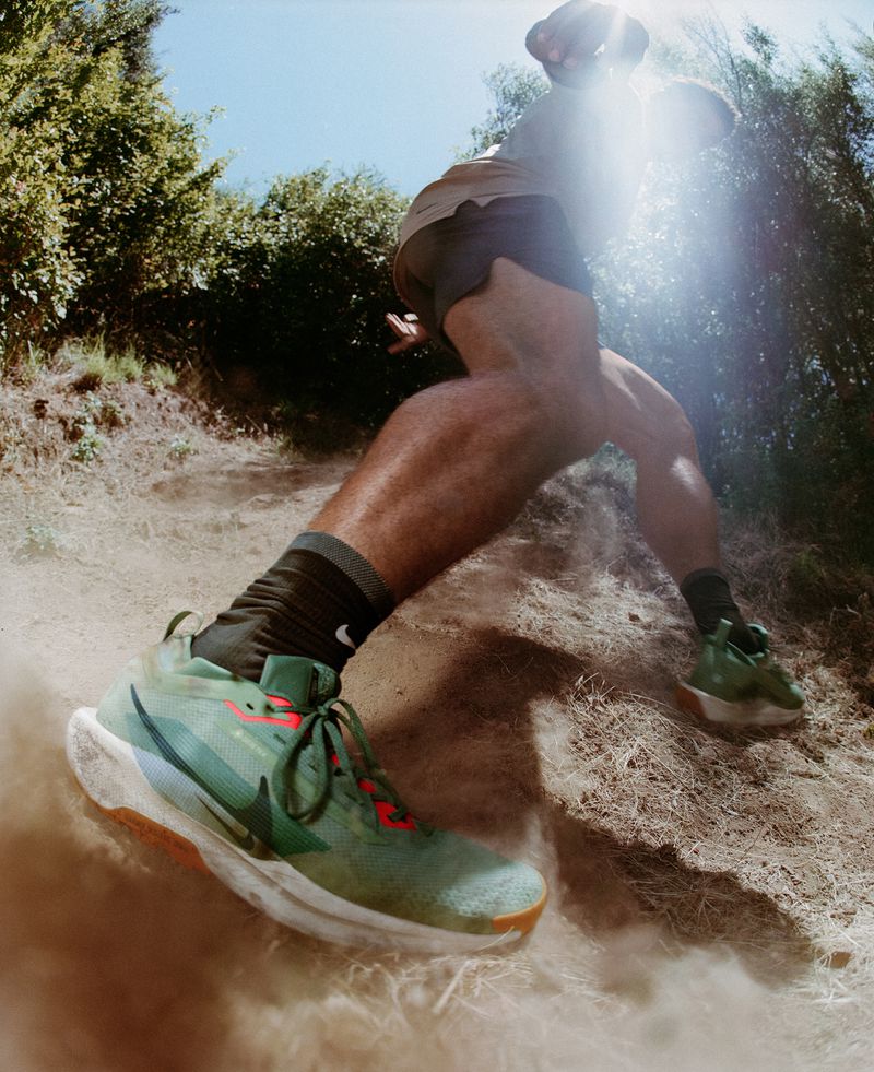 Athletic man running outdoors on a dirt trail, wearing shorts and athletic shoes. Ground level, eye-level perspective with bright sunlight.
