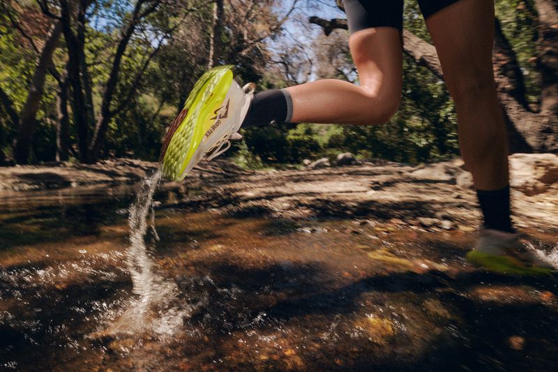 An adult athlete splashes through a stream in running shoes, in motion, with lush green trees visible in the background.