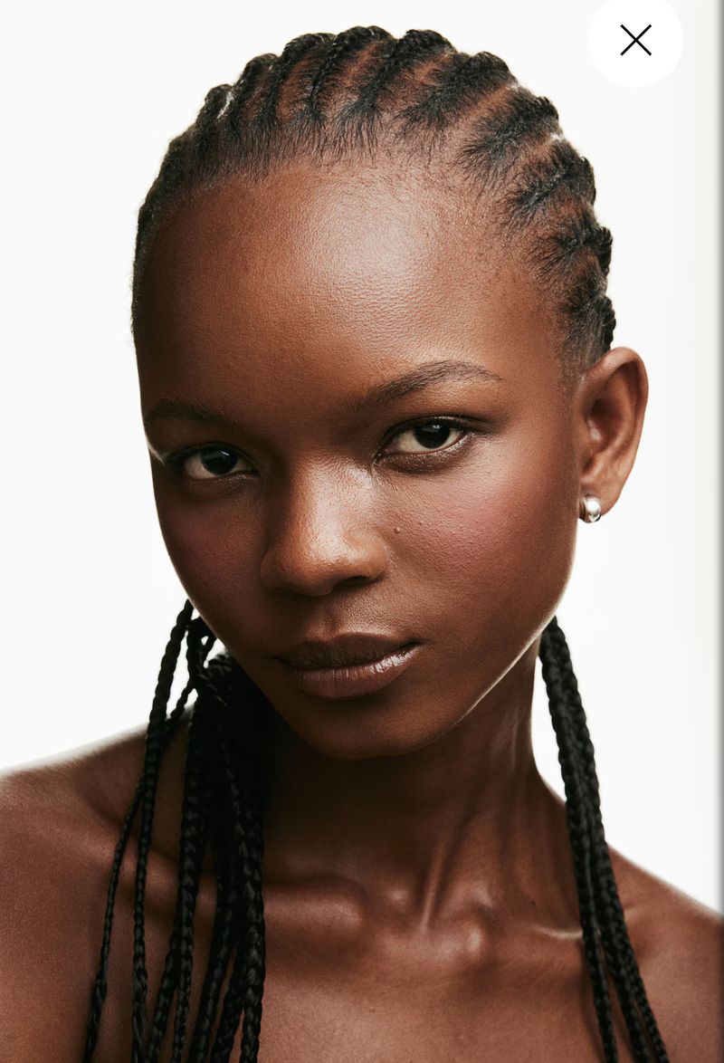 Close-up portrait of an African-descent woman with cornrow braids hairstyle, wearing a small pearl earring, set against a clean white backdrop.