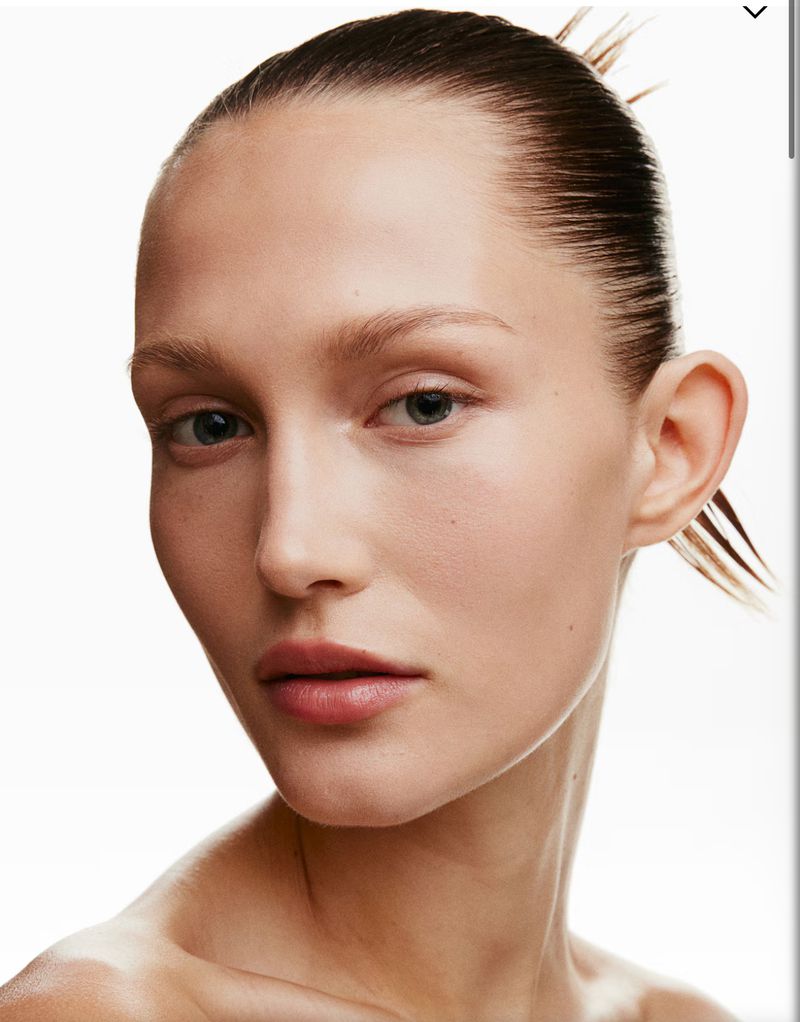 Close-up portrait of an adult female model with slicked-back hair and bare shoulders, set against a bright white background, emphasizing clean beauty.