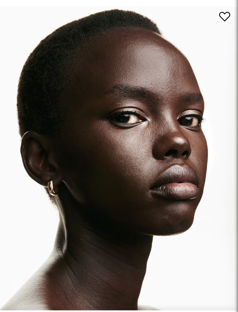 Close-up portrait of an adult woman with dark skin and short black hair against a white background, highlighting her facial features and gold earrings.