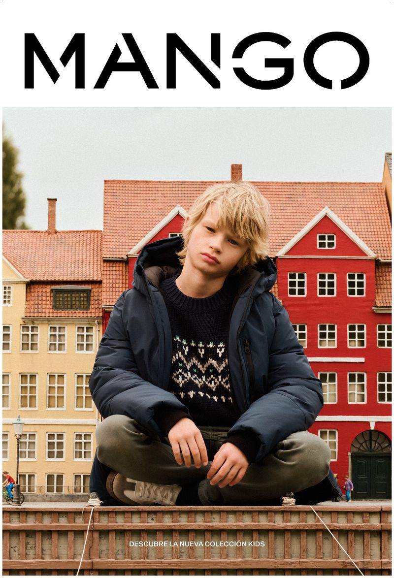 A young boy models Mango Kids' outerwear in an outdoor setting with red and yellow buildings; he is wearing a coat and patterned sweater.