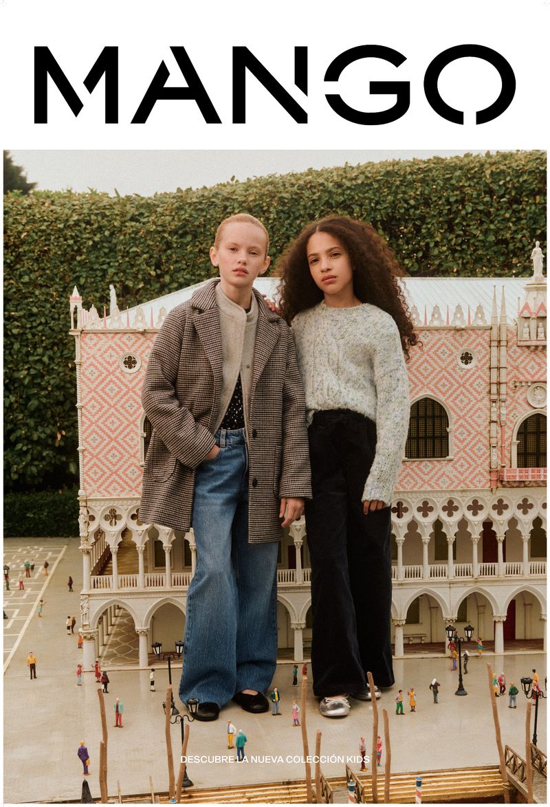 Two young girls in a Mango kids fashion ad, wearing coats and jeans, standing in front of a miniature Italian-style building outdoors.