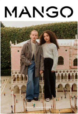 Two young girls in a Mango kids fashion ad, wearing coats and jeans, standing in front of a miniature Italian-style building outdoors.