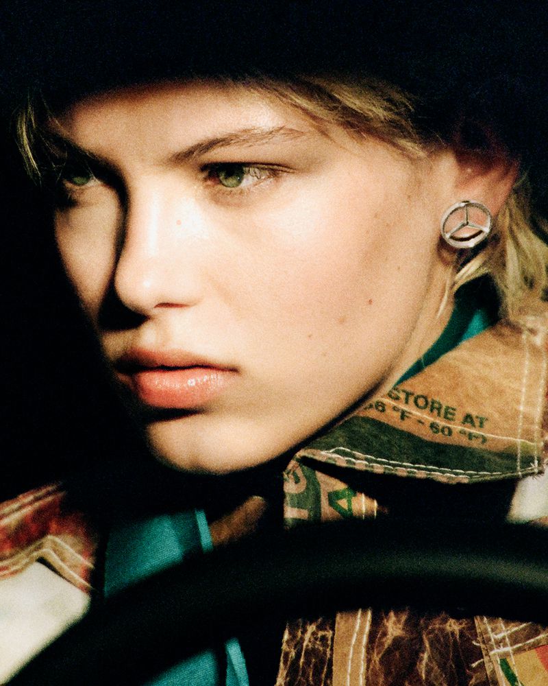 Close-up of a young adult woman with light skin wearing a fur hat and patterned jacket, looking off-camera with a serious expression in dim lighting.