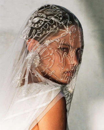 Close-up portrait of a woman wearing a white sheer embroidered veil and dress in a light studio setting, soft, diffused lighting.