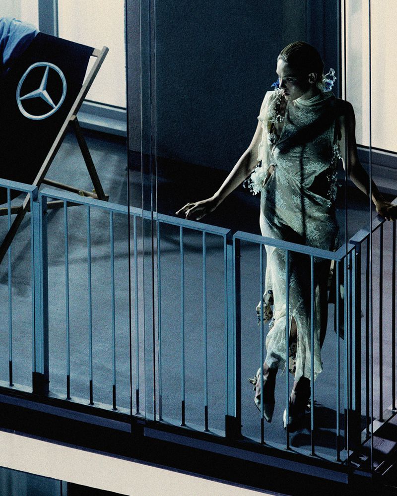 An adult female in a sheer dress poses on a balcony with a railing, alongside a beach chair displaying the Mercedes-Benz logo.