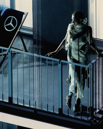 An adult female in a sheer dress poses on a balcony with a railing, alongside a beach chair displaying the Mercedes-Benz logo.