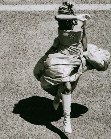 Woman in dress and boots standing in grass field with shadow, overhead view. Fashion shoot with automotive logo on dress.