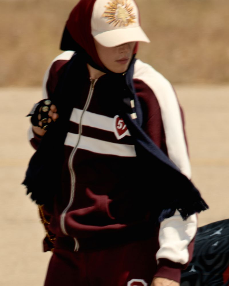 Stylish person in a maroon and white tracksuit with a cap poses outdoors, showcasing fashion and urban style against a neutral backdrop.