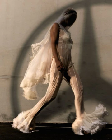 An adult woman is walking toward the left, wearing a sheer white jumpsuit with lace trim, a matching head covering, and a shadow behind her.