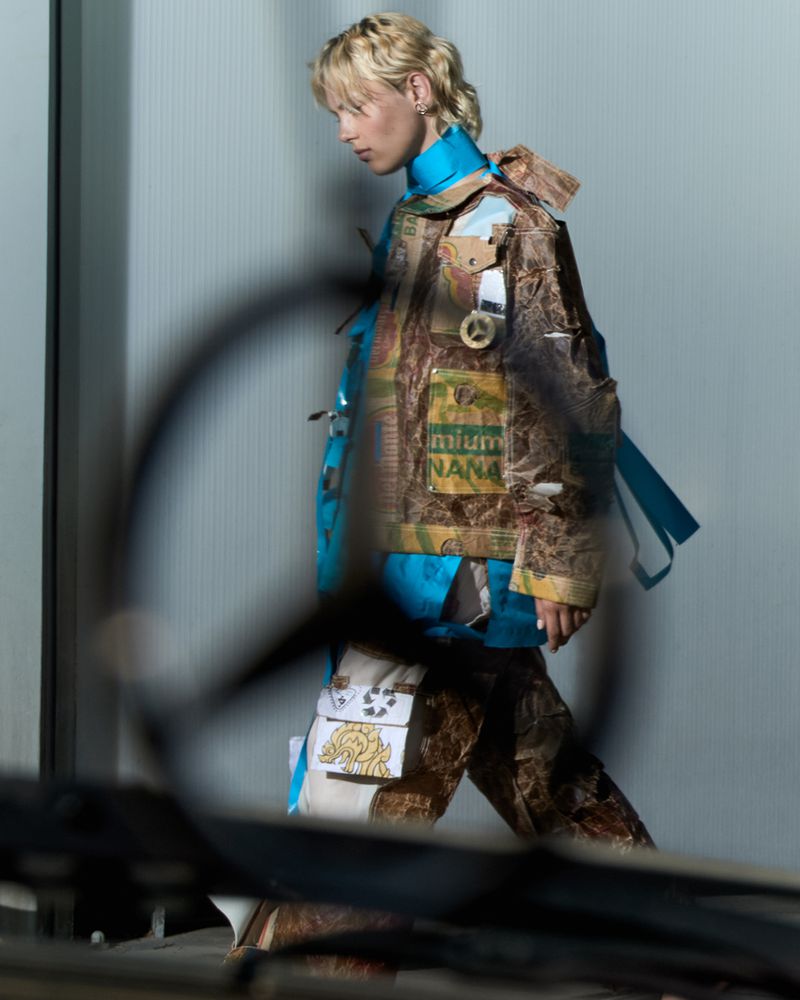A person with blonde hair wearing upcycled fashion strides against a grey wall, partially obscured by a blurred foreground element.