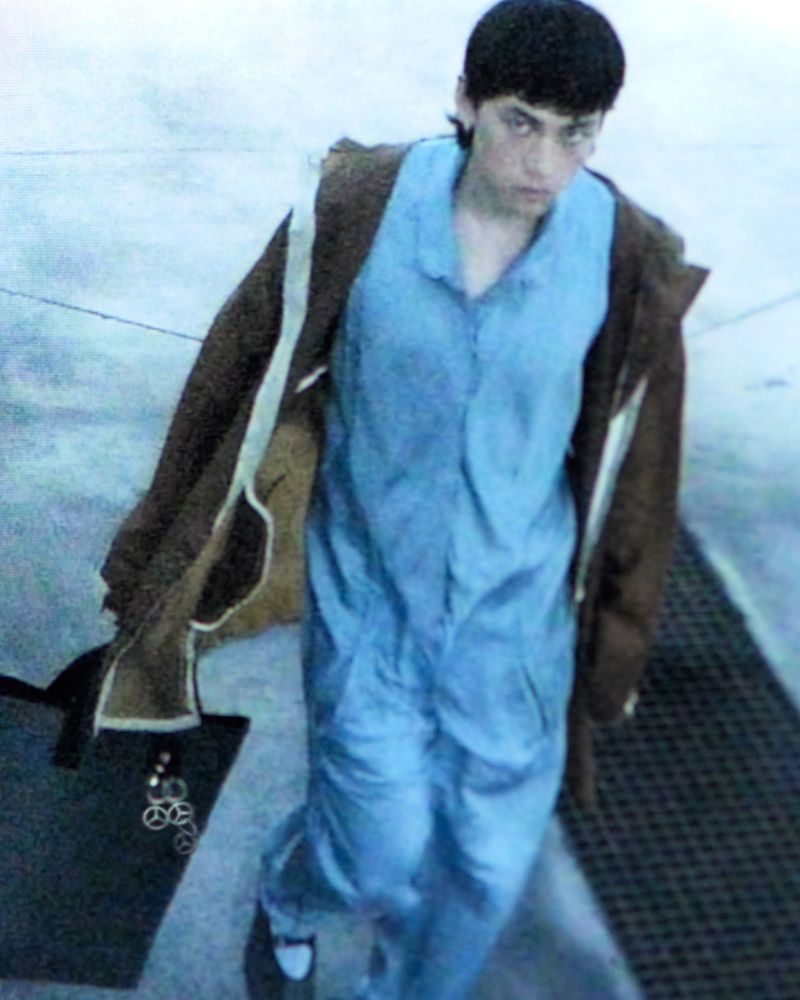 A young adult poses in a blue jumpsuit and brown jacket in an outdoor setting with urban textures, captured from a high angle.