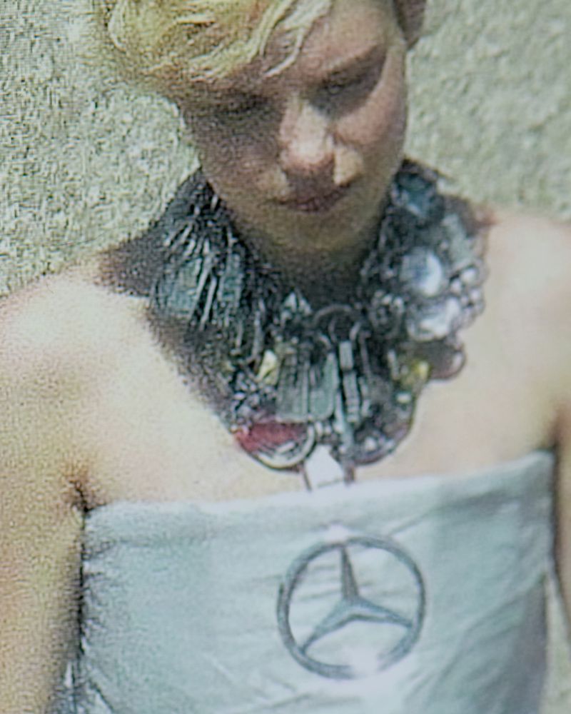 Woman in strapless dress with Mercedes-Benz logo and upcycled metallic trash necklace, stands against rough, sunlit outdoor backdrop.