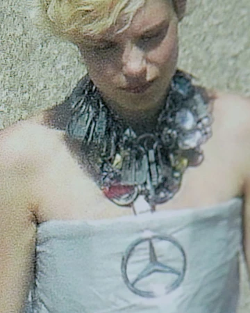 Woman in strapless dress with Mercedes-Benz logo and upcycled metallic trash necklace, stands against rough, sunlit outdoor backdrop.