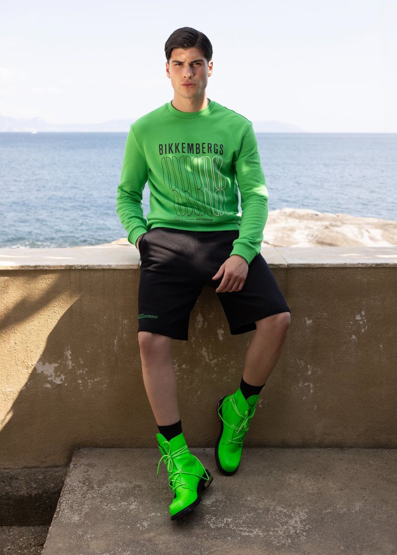 Young man poses in green sweatshirt, black shorts, and neon boots against an outdoor coastal backdrop on a sunny day.