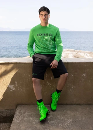 Young man poses in green sweatshirt, black shorts, and neon boots against an outdoor coastal backdrop on a sunny day.