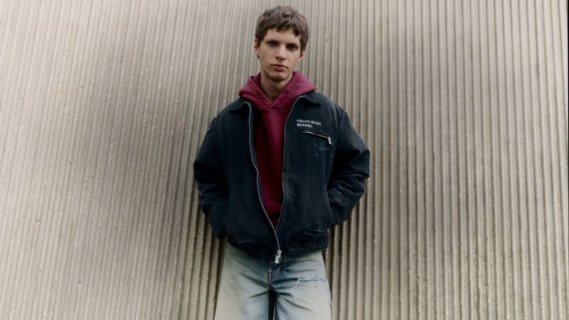 A person stands outdoors wearing a jacket and hoodie. The outfit is complemented by a textured wall backdrop enhancing the fashion-forward aesthetic.