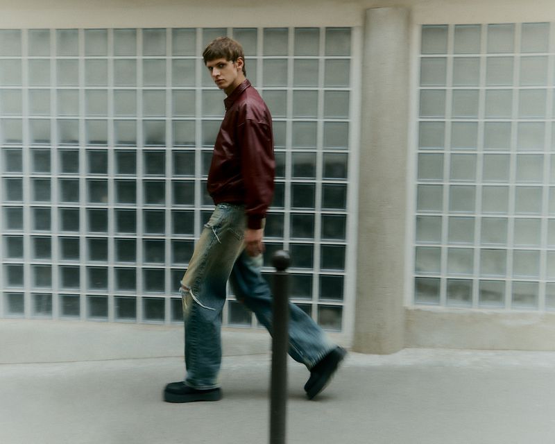 A male fashion model walks in ripped jeans, a burgundy jacket and black platform shoes near a glass block wall and street bollard.