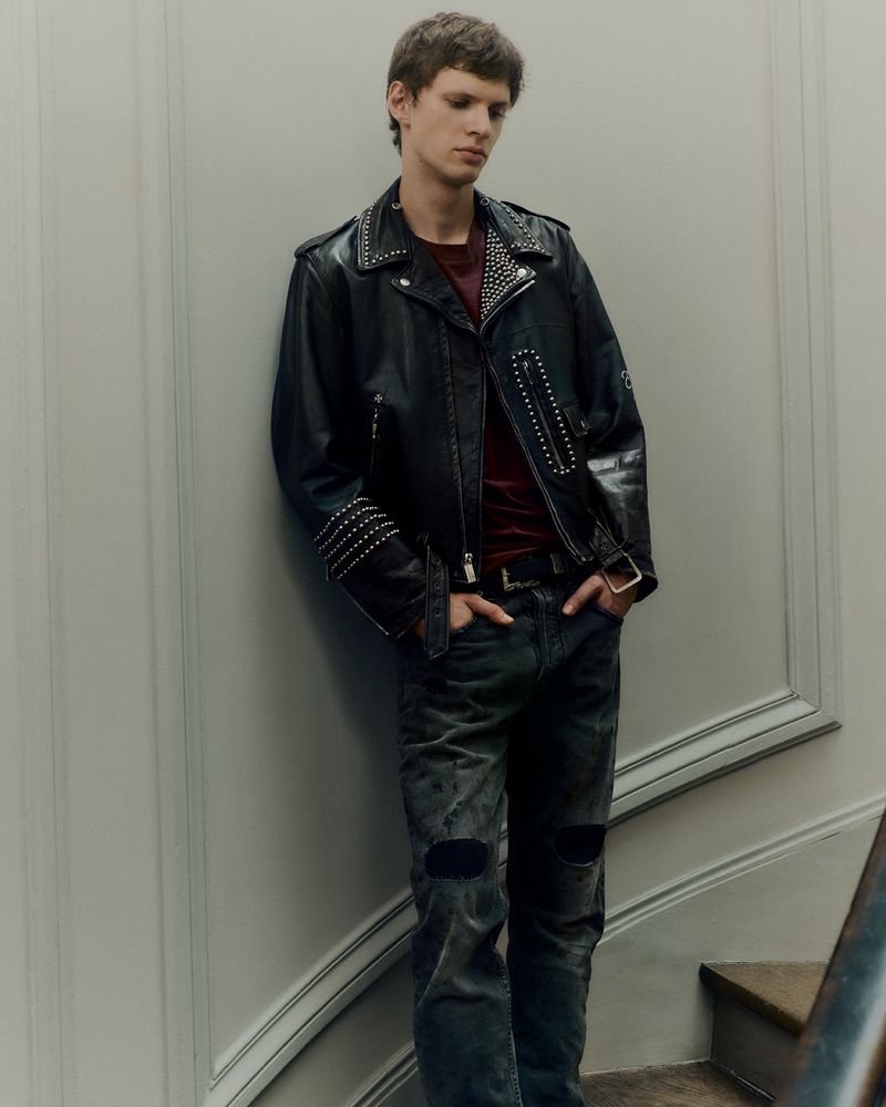Man in black studded leather jacket and ripped jeans leaning against a wall and staircase. Fashion editorial portrait.