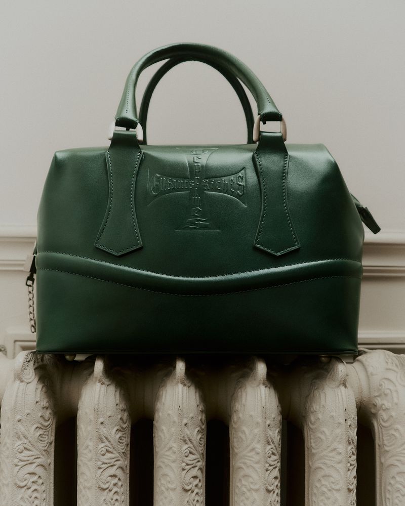 Green leather handbag with top handles and debossed logo detail on a white radiator with cream-colored wall in the background. Fashion.