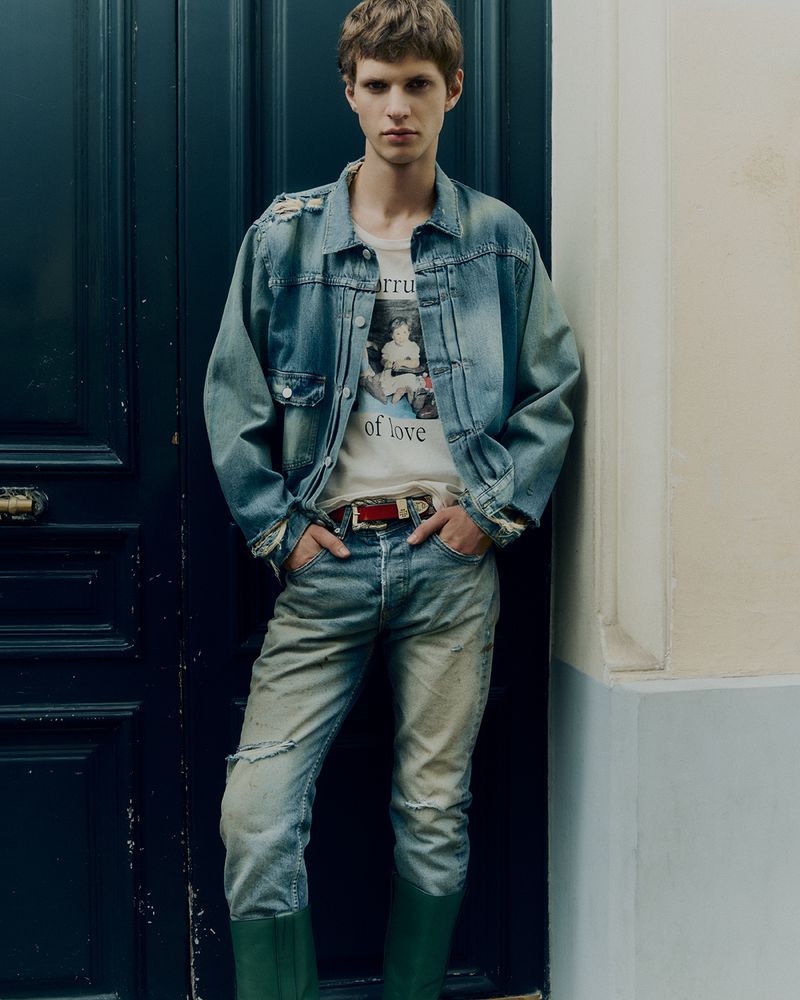 Male model in a distressed denim jacket, graphic tee, and jeans, posing against a dark blue door, fashion editorial shot.