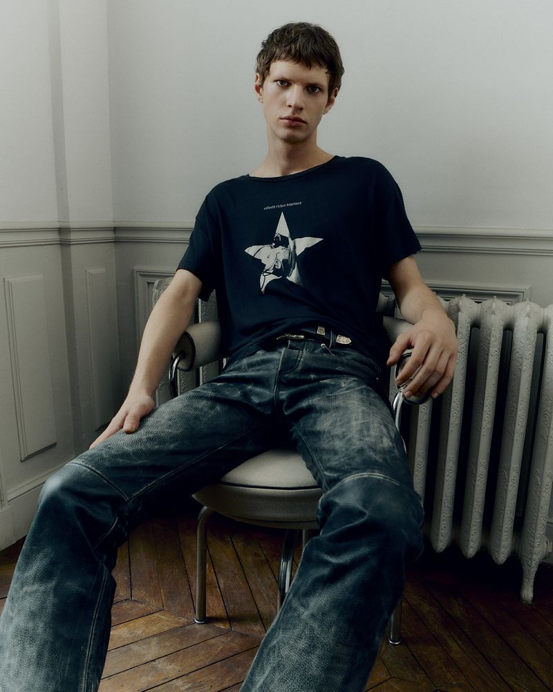 A young adult male with short brown hair sits on a chair wearing a printed t-shirt and distressed jeans inside a room with moulding.