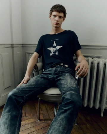 A young adult male with short brown hair sits on a chair wearing a printed t-shirt and distressed jeans inside a room with moulding.