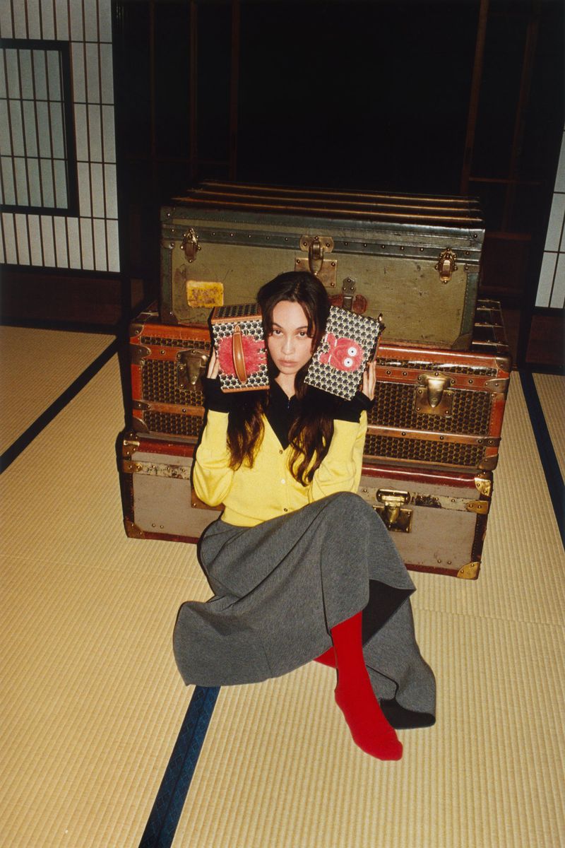 Woman in yellow cardigan sits with vintage luggage trunks, holding patterned cases, wearing red socks on tatami mat indoors.
