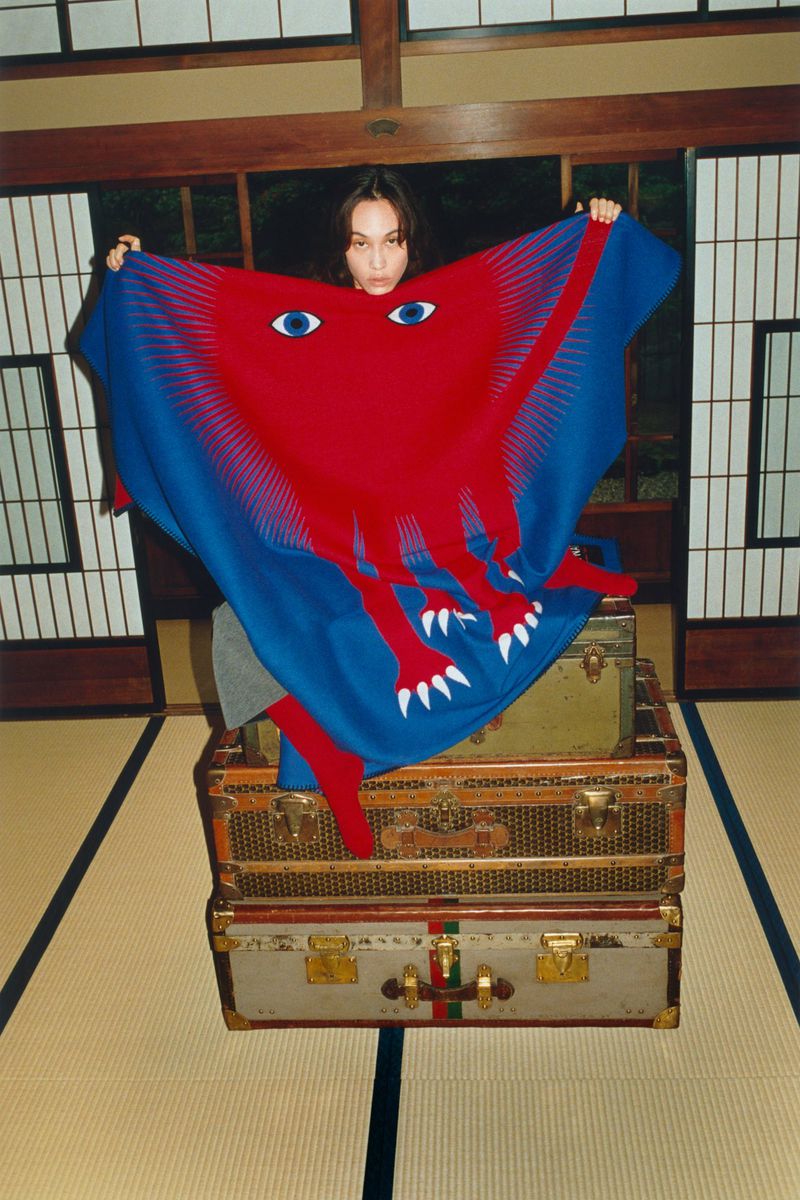 Woman in interior holding blue and red textile with cartoon eyes, seated on vintage suitcases against traditional Japanese shoji screen.