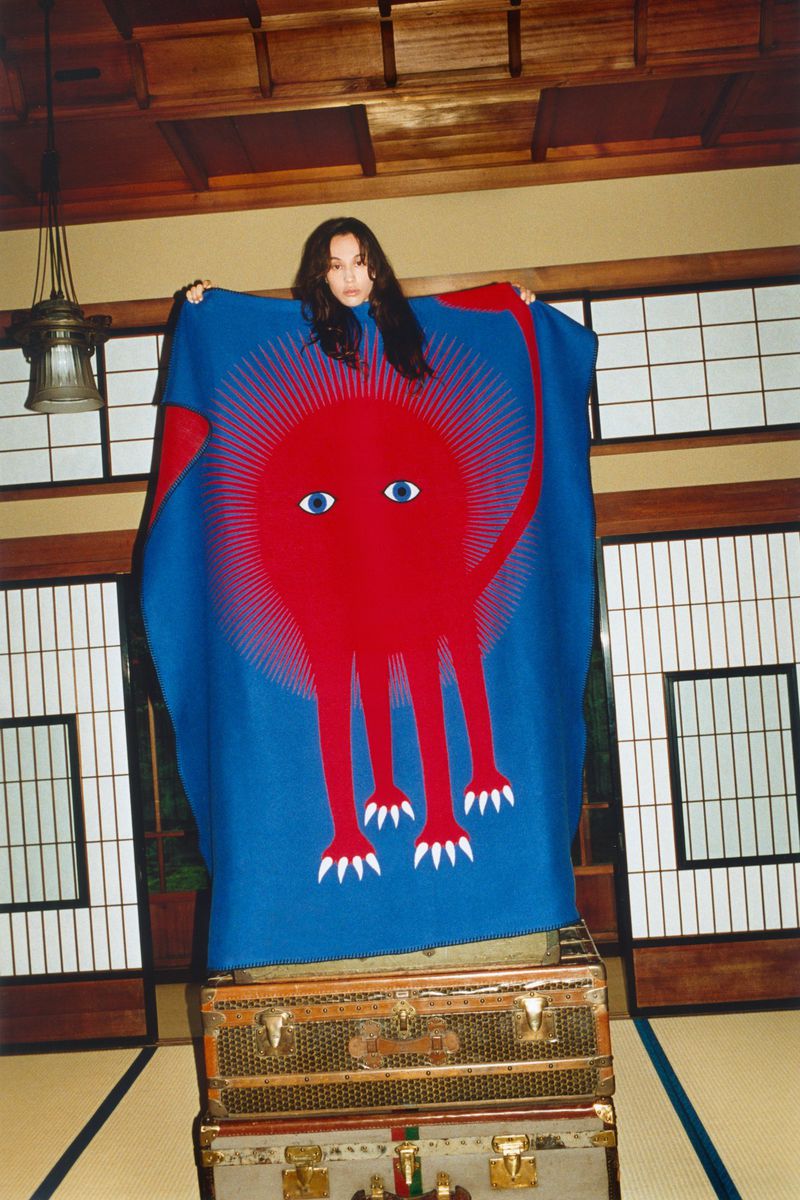 A woman stands behind a blanket with a red monster graphic, posed in a traditional Japanese room, on top of stacked vintage suitcases.