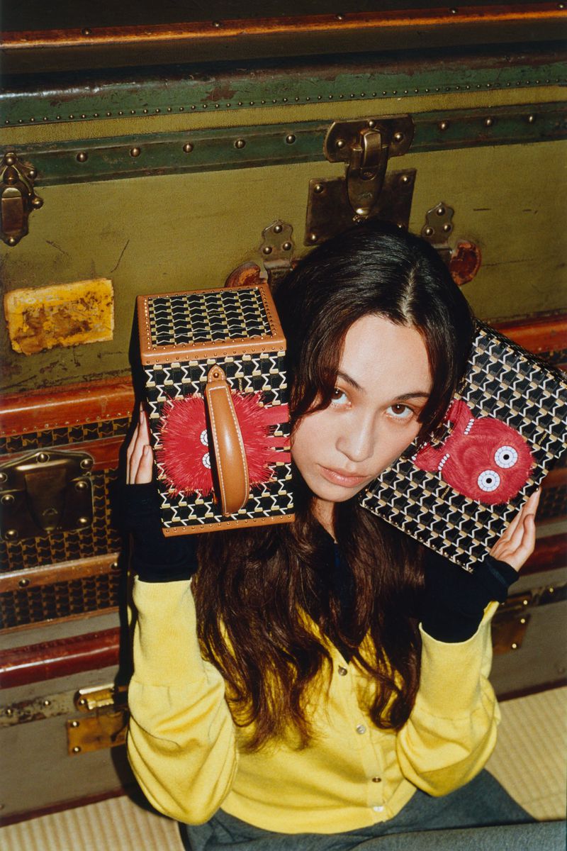 Portrait of a young adult woman holding two patterned handbags with red plush appliqués, posing indoors against a backdrop of vintage trunks.
