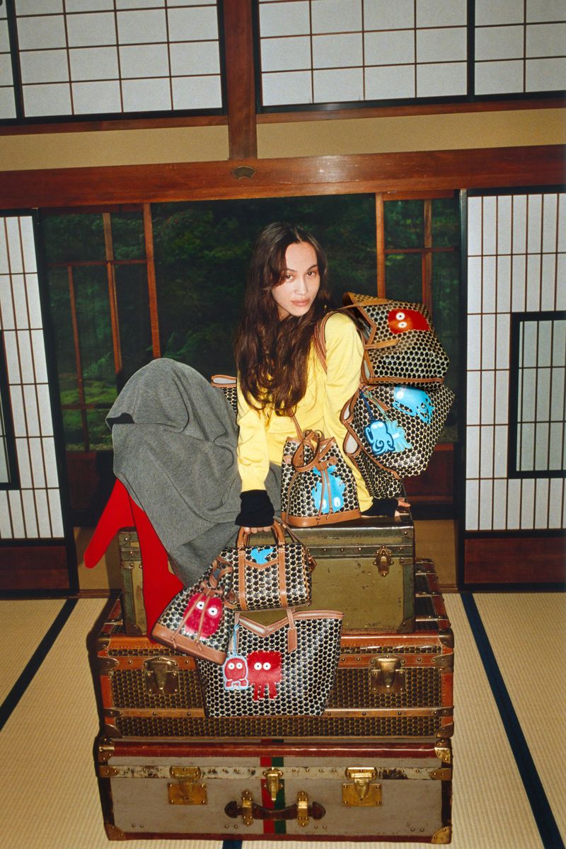 A woman poses in a yellow sweater next to several designer bags on top of vintage trunks inside a traditional Japanese room.