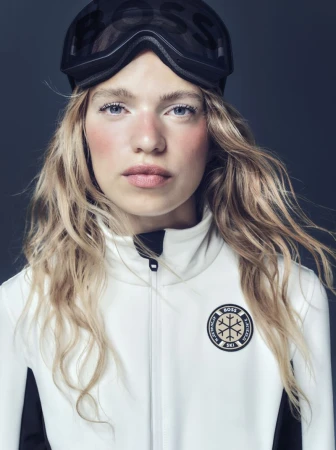 Close-up of a blonde woman in a white ski jacket and ski goggles on her head against a grey background in a studio setting.