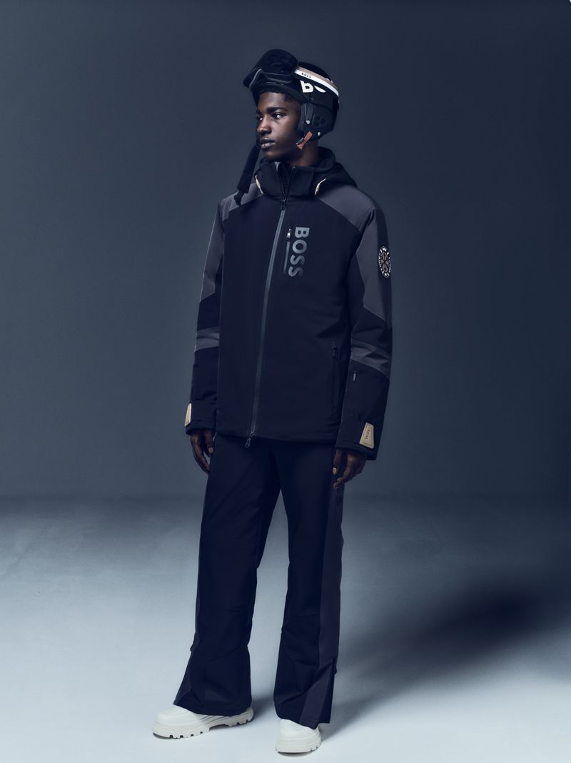 Male model poses in black BOSS ski jacket, ski pants, helmet, and snow boots against a grey gradient backdrop in a studio shot.